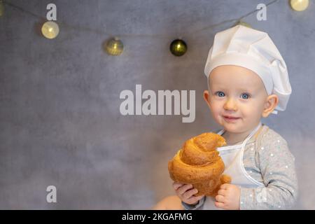 una ragazza carina in un cappello da chef e un grembiule siede a casa sul tavolo in cucina con un croissant Foto Stock