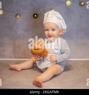 una ragazza carina in un cappello da chef e un grembiule siede a casa sul tavolo in cucina con un croissant Foto Stock