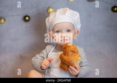 una ragazza carina in un cappello da chef e un grembiule siede a casa sul tavolo in cucina con un croissant Foto Stock