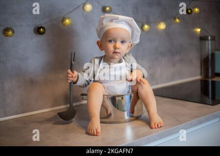 un bambino piccolo e carino si siede in cucina sul tavolo in una pentola con un mestolo nel cappello e grembiule di uno chef. Foto Stock