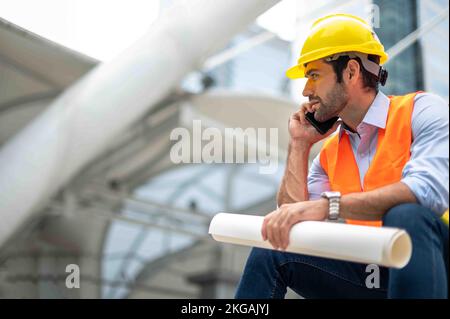 Gli ingegneri dell'uomo caucasici usano uno smartphone per parlare, indossando un giubbotto arancione e un grande elmetto, e l'altra mano che tiene la pianta bianca del sito Foto Stock