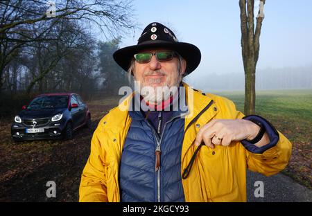 Itterbeck, Germania - Febbraio 28 2021 Un uomo con un impermeabile giallo e un cappello nero da cowboy con spille davanti ad un'auto elettrica nera: Un EQ Smart forfour Foto Stock