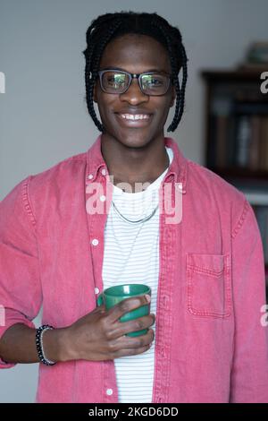 Felice uomo afro-americano positivo si alza in casa con tazza in mano e sorridente guarda la macchina fotografica Foto Stock