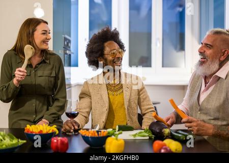 Gruppo di amici vegetariani che si divertono a cena, cucinando cibi crudi e verdure, bevendo vino, gente mista anziana e multirazziale che scherza al celebrato Foto Stock