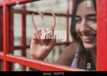 Donna, cuore e disegno con rossetto in stand per amore in vacanza di viaggio a Londra città. Trucco di cosmetici rossi e graffiti della persona felice sulla finestra Foto Stock
