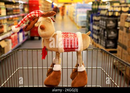 Natale, Capodanno, shopping. Capriolo di Natale divertente sul carrello del supermercato. Paniere in negozio all'ingrosso, sfondo di alimentari sfocati e merci. Foto Stock