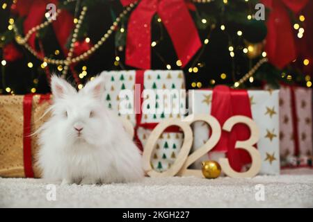 Vista frontale di un grazioso coniglio peloso seduto su un tappeto bianco guardando la macchina fotografica. Simbolo di nuovo anno che ha fotografato vicino all'albero decorato di natale. Conc Foto Stock