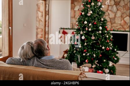 Coppia anziana, natale e abbraccio dalla vista posteriore rilassarsi sul divano nel soggiorno con albero di natale. Amore, sostegno e festa di festa o Foto Stock
