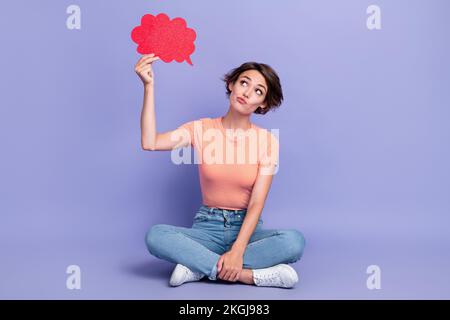 Foto a corpo pieno di bella giovane donna guardare incerto tenere fumetti discorso bolla indossare abito a righe trendy isolato su sfondo di colore viola Foto Stock