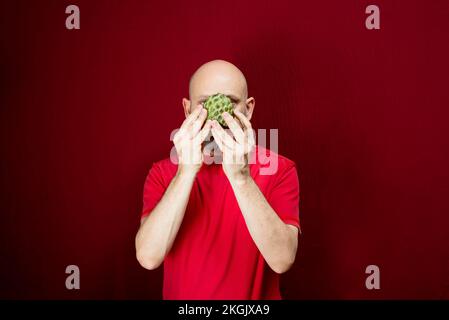 Giovane uomo bello con barba, testa calva e camicia rossa in piedi che tiene la frutta di pinecone davanti al viso contro sfondo rosso. Foto Stock