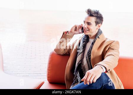 Un giovane uomo caucasico seduto all'aperto sulla panchina il giorno d'autunno tenendo il telefono cellulare e parlando Foto Stock