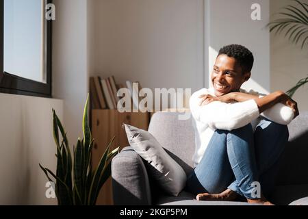Donna sorridente e rilassata seduta sul divano a casa guardando fuori dalla finestra Foto Stock