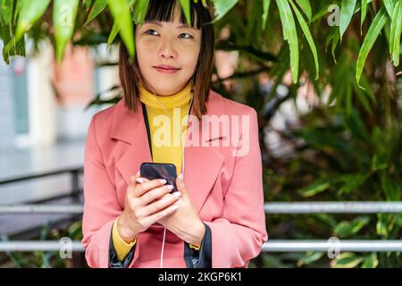 Giovane donna sorridente in piedi con smartphone e ascoltare musica attraverso le cuffie al parco Foto Stock