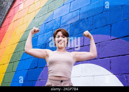 Giovane donna felice che flette i muscoli di fronte alla bandiera arcobaleno dipinta sulla parete Foto Stock