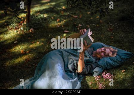 Donna matura lettura libro sdraiato in giardino Foto Stock