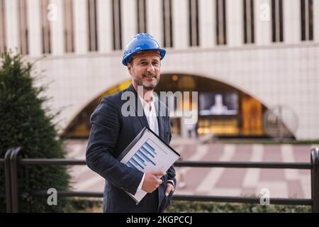 Uomo d'affari felice che indossa un hardHat che tiene la carta di fronte alla ringhiera Foto Stock