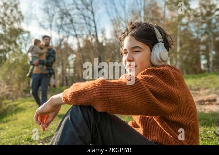 Ragazzo che ascolta la musica tramite cuffie wireless con un uomo che porta il figlio in sottofondo Foto Stock