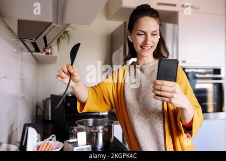 Giovane donna sorridente che tiene il cucchiaio di legno che ha videochiamata attraverso il telefono cellulare in cucina Foto Stock