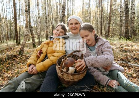 Nonna sorridente, ragazzo e ragazza seduti con cestino di funghi nella foresta Foto Stock