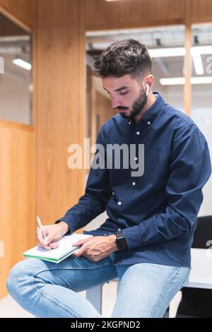 Giovane uomo d'affari che scrive in agenda seduto sulla scrivania in ufficio Foto Stock