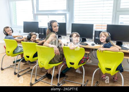 Sorridendo gli studenti con un insegnante che indossa delle cuffie seduto a scuola in una lezione di computer Foto Stock