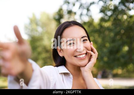 Sorridente ragazza asiatica prende selfie, video chat, tiene lo smartphone e parla alla fotocamera, in posa in parco Foto Stock