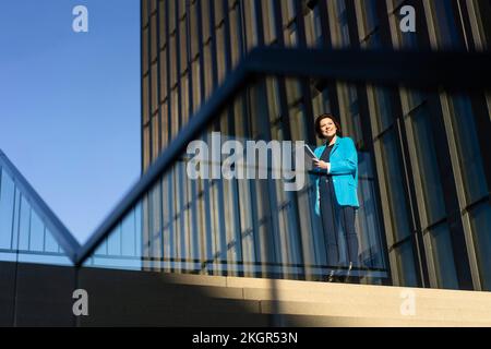 Donna d'affari sorridente in piedi vicino all'edificio visto attraverso la ringhiera di vetro Foto Stock