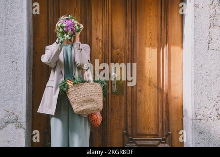 Donna che tiene borse coprire il viso con cavolfiore viola di fronte alla porta Foto Stock