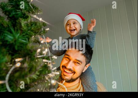 Sorridente ragazzo seduto sulla spalla del padre che ha divertente decorare l'albero di Natale a casa Foto Stock