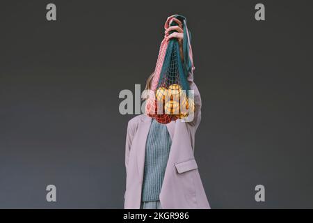 Donna che copre il viso con una borsa a rete di fronte alla parete grigia Foto Stock
