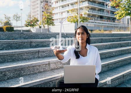 Giovane donna con modello a turbina eolica e computer portatile seduto sui gradini Foto Stock