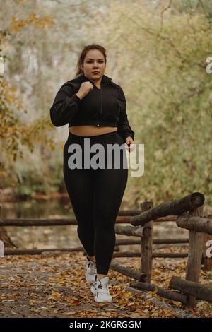 Una giovane donna in sovrappeso determinata che fa jogging nel parco autunnale Foto Stock