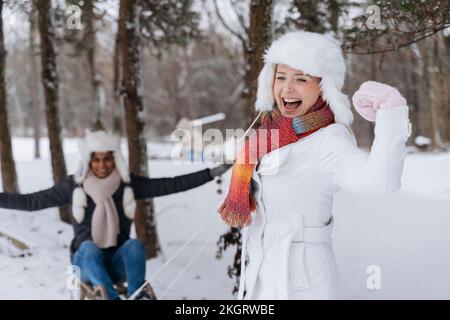 Donna felice che flette i muscoli con l'uomo seduto sulla slitta nella neve Foto Stock