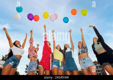 Amici con le mani sollevate tenendo palloncini colorati sotto il cielo blu Foto Stock