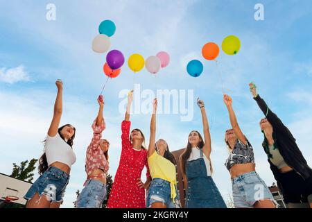 Amici felici con le mani sollevate tenendo palloncini colorati sotto il cielo blu Foto Stock