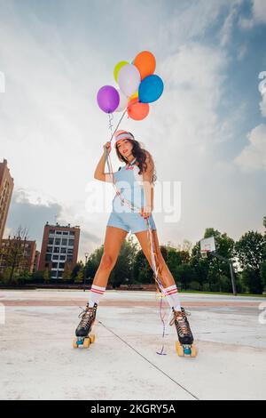 Giovane donna che indossa pattini a rotelle con palloncini colorati sotto il cielo a campo sportivo Foto Stock