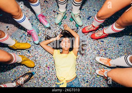 Donna felice sdraiata sui coriandoli fra le gambe degli amici che indossano pattini a rotelle Foto Stock