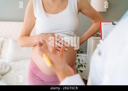 Donna incinta con mano sull'anca che tocca la pancia di fronte al medico a casa Foto Stock