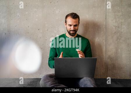 Uomo d'affari che effettua videochiamate tramite un computer portatile seduto davanti alla parete dell'ufficio Foto Stock