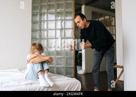 In piedi giovane padre aggressivo che scola, alzando la voce, gridando a bambino difficile e testardo depresso che siede sul letto che abbraccia il ginocchio. Foto Stock