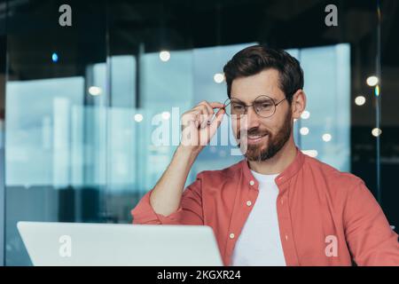 Foto ravvicinata di un architetto di successo e felice che lavora all'interno di uno studio di design moderno, uomo in occhiali e camicia rossa sorridente e che guarda lo schermo del notebook. Foto Stock