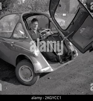 1960s, storico, fuori in un parcheggio di ghiaia, un uomo seduto all'interno di una piccola auto dell'epoca, una bolla auto, una BMW Isetta, che aveva una porta di apertura anteriore, Wycombe, Inghilterra, Regno Unito. Costruita per la prima volta da ISO Spa in Italia nel 1953, questa piccola auto è stata costruita su licenza in diversi paesi, tra cui BMW di Germania fino al 1962 e nota come la 'Bubble Car'. Foto Stock