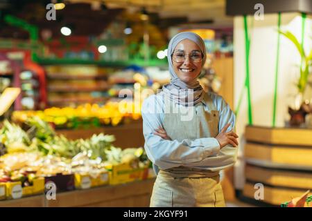 Ritratto donna in un hijab come un venditore in un supermercato negozio, un venditore con le braccia incrociate sorrisi e guarda la macchina fotografica, un grembiule vende mele e frutta, una donna musulmana è soddisfatto in bicchieri. Foto Stock