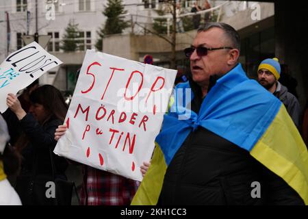 Manifestazione contro la guerra in Ucraina. Un uomo ha una bandiera Ucraina intorno alla sua spalla. Qualcuno ha un segno: "Stop assassin Putin”. Foto Stock