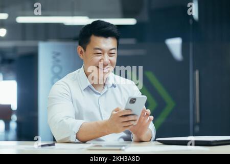 Giovane bell'uomo asiatico, lavoratore seduto in ufficio al tavolo, cullare con il telefono, scrivere messaggi, chiacchierare. Foto Stock