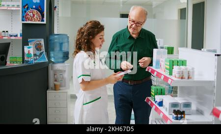 Dipendente della farmacia che fornisce assistenza a clienti diversi, aiutando i clienti a scegliere il trattamento sanitario o integratori. Farmacista che spiega medicamenti e prodotti sugli scaffali. Foto Stock