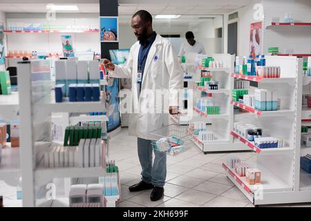 Farmacista afroamericano che controlla i supplementi nutrizionali in scaffale della farmacia, tenendo il cestino di shopping pieno dei prodotti farmaceutici. Vendita al dettaglio di farmaci, articoli farmaceutici Foto Stock