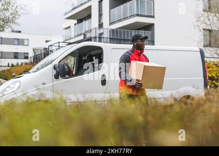 Lavoratore con colletto blu nero - addetto alla consegna dell'imballaggio - davanti al camion bianco con imballaggio in cartone. Abbigliamento - giacca rossa con maniche grigie e cappello nero. Erba alta sfocata in primo piano. Foto di alta qualità Foto Stock