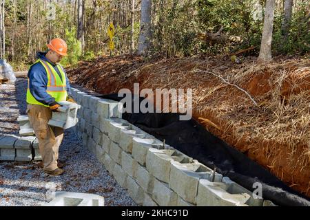 L'appaltatore che installa la parete del blocco di calcestruzzo come parte della costruzione della parete di ritegno che è in fase di costruzione come parte del progetto Foto Stock