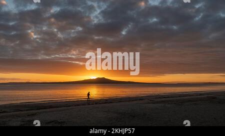 Silhouette donna che cammina sulla spiaggia di Milford. Sole che sorge sull'Isola di Rangitoto. Auckland. Foto Stock